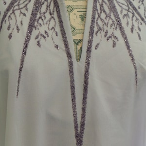 May include: A light grey blouse with a V-neckline and a decorative design of purple sequins in the shape of branches and leaves.