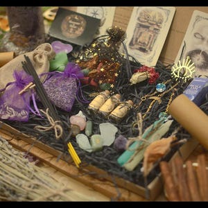 May include: A wooden box containing spiritual items such as purple pouches, incense sticks, crystals, small bottles, dried orange slices, and rolled scrolls. The box is decorated with dried leaves and other natural elements.