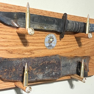 May include: A wooden display board featuring a fixed-blade knife and a leather sheath mounted on four brass bullet-shaped pegs. A circular U.S. Marine Corps emblem is centered between the knife and sheath, with engraved text on the leather.