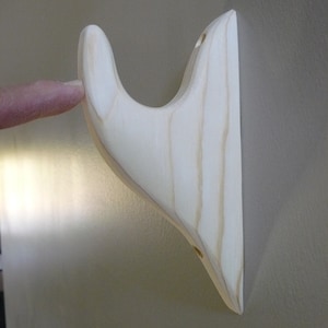 May include: A light wood wall mounted hook with a curved design. The hook is unfinished and has two holes for mounting.