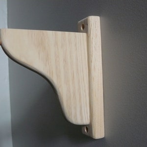 May include: A wooden shelf bracket with a curved edge and two holes for mounting. The bracket is made of unfinished wood and is ready to be stained or painted.