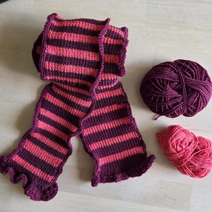 Può includere: Una sciarpa lavorata a maglia con strisce orizzontali viola scuro e rosa corallo con bordi arricciati, accompagnata da un gomitolo di lana viola scuro e uno rosa corallo, che mostrano la consistenza e la tavolozza dei colori dell'articolo.