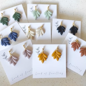 Puede incluir: Una colección de pendientes con forma de hoja en varios colores, incluyendo verde, azul, crema, negro, malva, amarillo mostaza y óxido. Cada par se exhibe en una tarjeta blanca con un poste dorado. Las tarjetas tienen un diseño de hoja y el texto "Land of Jewellery."