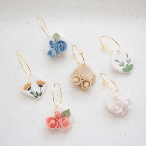 Può includere: Sei paia di orecchini in ceramica fatti a mano con motivi floreali. Gli orecchini presentano una varietà di fiori, tra cui rose, margherite e foglie. Gli orecchini sono tutti in ceramica bianca e hanno cerchi dorati.