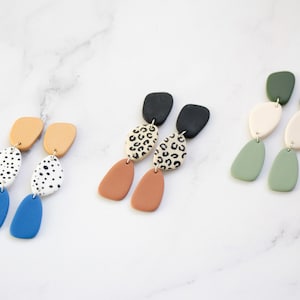 May include: Three pairs of dangle earrings with geometric shapes. The earrings are made of polymer clay and feature different patterns, including polka dots, leopard print, and solid colors. The earrings are in shades of blue, green, brown, white, and black.