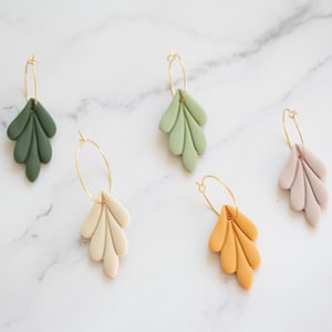 May include: Four pairs of gold hoop earrings with dangling leaf-shaped charms. The charms are made of polymer clay and come in four colors: green, cream, light brown, and light purple.