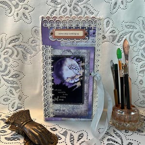 Puede incluir: Un diario hecho a mano con un diseño morado y blanco. La portada presenta una ilustración de luna y mariposa, con el texto "never stop looking up" y "tell your secrets to the rising moon". El diario está encuadernado con encaje blanco y una cinta azul claro.