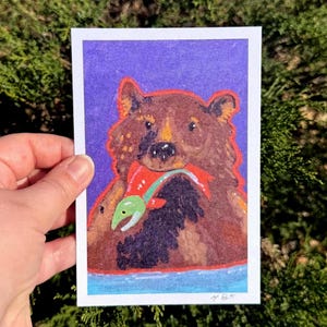 May include: A colorful illustration of a brown bear holding a red fish with a green stripe against a purple background. The bear is depicted with orange outlines, and the fish has a green head. The artwork is on a white rectangular card.