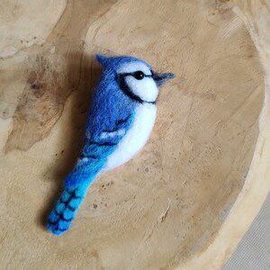 May include: A handcrafted blue jay bird figurine with white accents and a black beak. The bird is made of felted wool and is set against a light brown wooden background. The bird's feathers are detailed with darker blue markings.