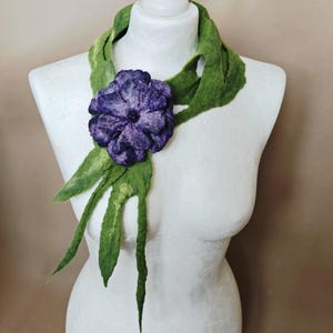 May include: A green felt scarf with a large, purple flower accent. The scarf is draped on a white mannequin. The flower is a deep purple with a dark center. The scarf has long, leaf-like extensions.