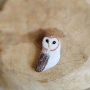 May include: A handmade felt owl figurine. The owl has a white body with brown and grey wing markings. It features a brown outline around the face and black eyes. The owl is a miniature decorative item.