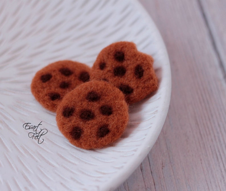 Pu&ograve; includere: Tre biscotti di feltro marroni con gocce di cioccolato su un piatto bianco. I biscotti sono fatti a mano e hanno una consistenza realistica.