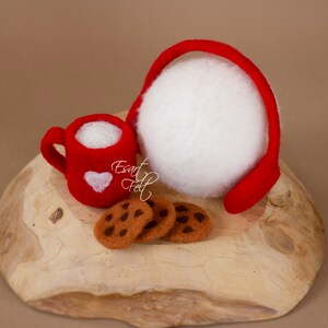 May include: A red felt mug with a white heart design, filled with white felt, next to three chocolate chip cookies on a wooden surface. A red felt headband with a white felt ball is also on the surface.