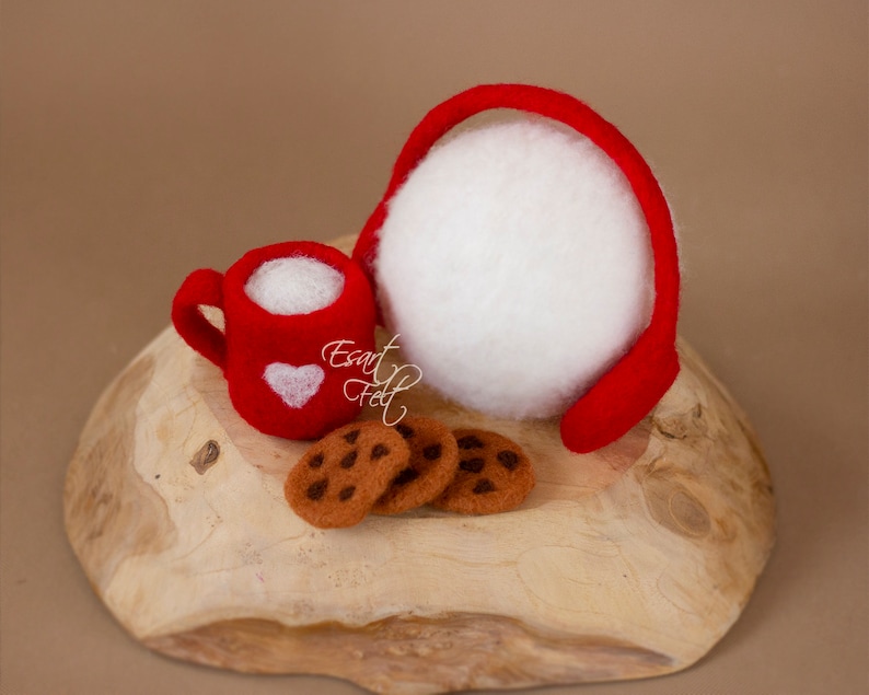 May include: A red felt mug with a white heart design, filled with white felt, next to three chocolate chip cookies on a wooden surface. A red felt headband with a white felt ball is also on the surface.