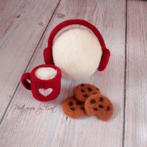 May include: Red felt mug with a white heart and a white felt ball with red felt earmuffs, along with three brown felt cookies on a white wood background.