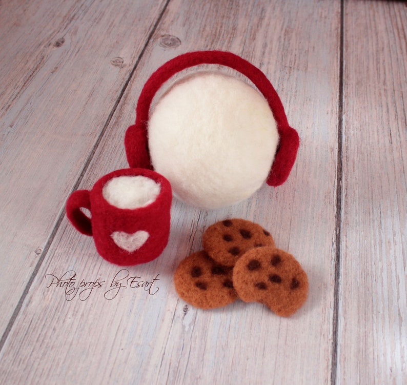 May include: Red felt mug with a white heart and a white felt ball with red felt earmuffs, along with three brown felt cookies on a white wood background.