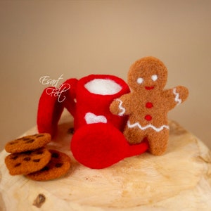 May include: A red felt mug with white hearts and a gingerbread man figurine, along with a stack of chocolate chip cookies on a wooden surface.