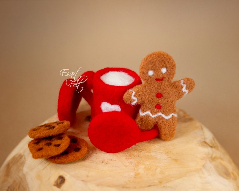 May include: A red felt mug with white hearts and a gingerbread man figurine, along with a stack of chocolate chip cookies on a wooden surface.