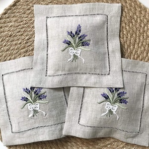 May include: Three square linen sachets with embroidered lavender sprigs and a white bow. The sachets are on a woven brown background.