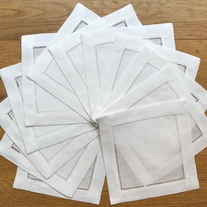 May include: A set of 12 white linen napkins with a delicate embroidered border. The napkins are arranged in a fan shape on a wooden surface.
