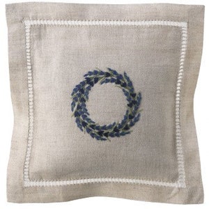 May include: A square linen napkin with a white embroidered border and a blue floral wreath design in the center.