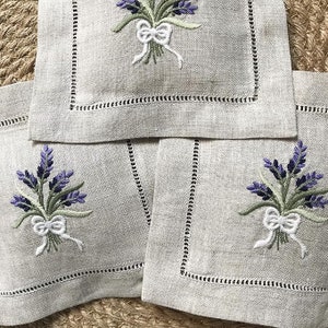 May include: Set of three linen napkins with embroidered lavender sprigs and white bows. The napkins are beige with a scalloped edge.