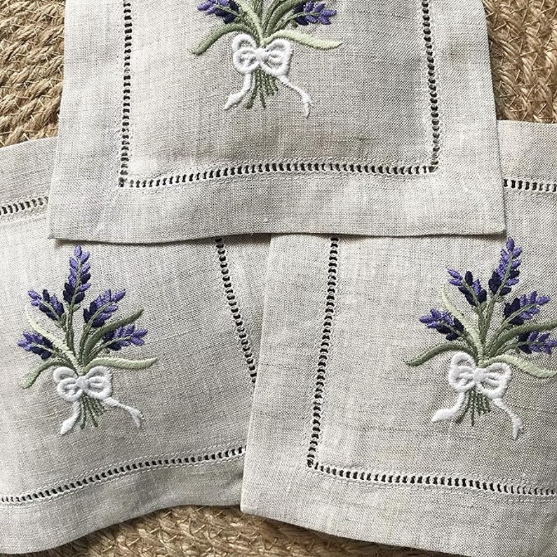 May include: Set of three linen napkins with embroidered lavender sprigs and white bows. The napkins are beige with a scalloped edge.