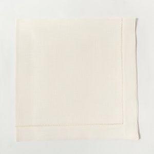 Puede incluir: Servilleta de lino color marfil con un borde cosido. La servilleta cuadrada est&aacute; doblada y colocada sobre una superficie blanca. La costura es visible a lo largo del borde de la servilleta, agregando un detalle decorativo. Esta elegante servilleta es adecuada para cenas formales.