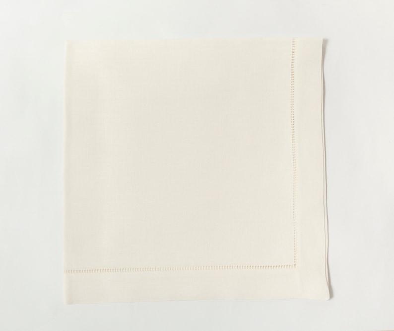 Puede incluir: Servilleta de lino color marfil con un borde cosido. La servilleta cuadrada est&aacute; doblada y colocada sobre una superficie blanca. La costura es visible a lo largo del borde de la servilleta, agregando un detalle decorativo. Esta elegante servilleta es adecuada para cenas formales.