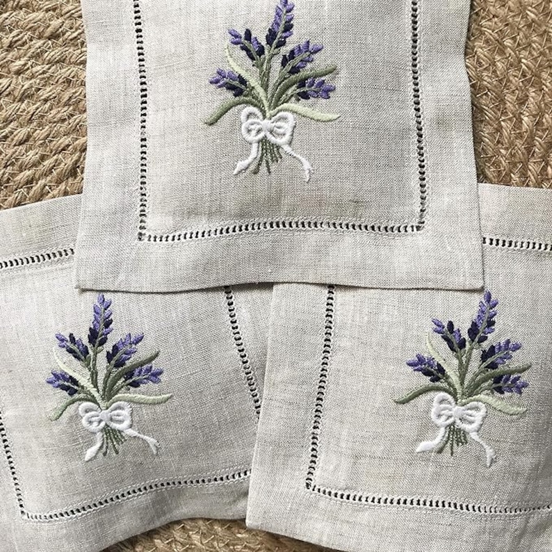 May include: Three linen napkins with embroidered lavender sprigs and white bows. The napkins are beige and have a decorative stitched border.