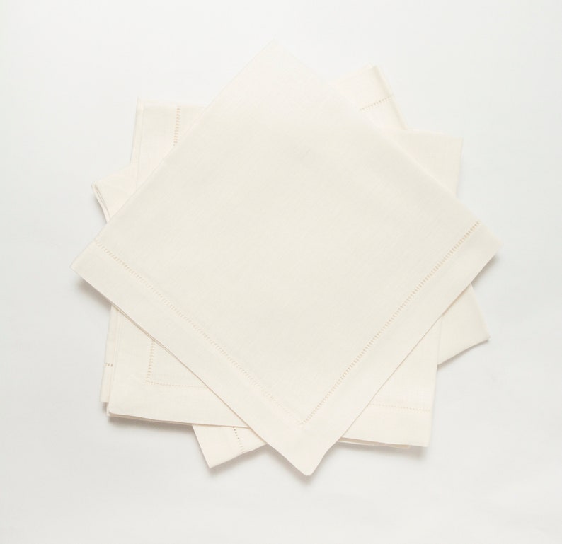 Puede incluir: Una pila de cuatro servilletas de lino blanquecino. Cada servilleta cuadrada tiene un borde cosido. Las servilletas est&aacute;n dispuestas de forma ligeramente superpuesta, mostrando su textura y dise&ntilde;o. El color neutro y el dise&ntilde;o sencillo sugieren un estilo cl&aacute;sico y elegante.