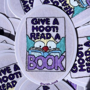 May include: A round sticker with a white background and a purple border. The sticker features a cartoon clown with a red nose and big eyes reading a book. The text "Give a Hoot! Read a Book" is written in purple letters above the clown.