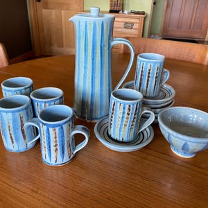Puede incluir: Un juego de tazas de cerámica rayadas en azul y marrón y una jarra a juego con tapa. Las tazas y la jarra tienen un diseño pintado a mano. También hay un pequeño cuenco de cerámica azul y marrón.
