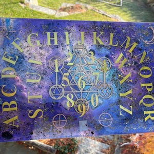 May include: A rectangular, purple and blue resin Ouija board with gold lettering and symbols. The board features the alphabet, numbers, and geometric shapes. Dried herbs and glitter are embedded in the resin.