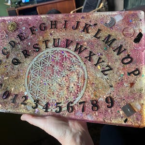 May include: A rectangular Ouija board with a pink and gold resin base. The board features black letters, numbers, and a decorative floral design in the center. Embellishments include gold charms and glitter.