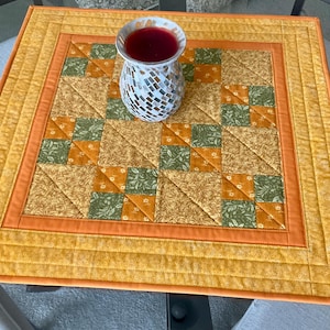 May include: A square, quilted table topper with a mosaic vase. The topper features a patchwork design in shades of orange, yellow, and green. The vase is white with a mosaic pattern and contains a red liquid.