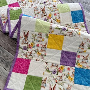 May include: Two quilted placemats with a bunny and floral print. The placemats feature a patchwork design with squares of white fabric with bunnies and flowers, and solid squares of pink, green, yellow, and blue. The edges are trimmed with a purple border.