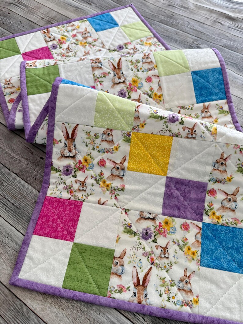 May include: Two quilted placemats with a bunny and floral print. The placemats feature a patchwork design with squares of white fabric with bunnies and flowers, and solid squares of pink, green, yellow, and blue. The edges are trimmed with a purple border.