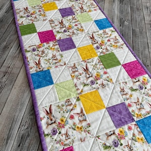 May include: A quilted Easter table runner featuring a repeating pattern of colorful squares and bunny illustrations. The squares are in shades of pink, yellow, green, purple, and blue. The runner has a purple border and is set on a wooden surface.