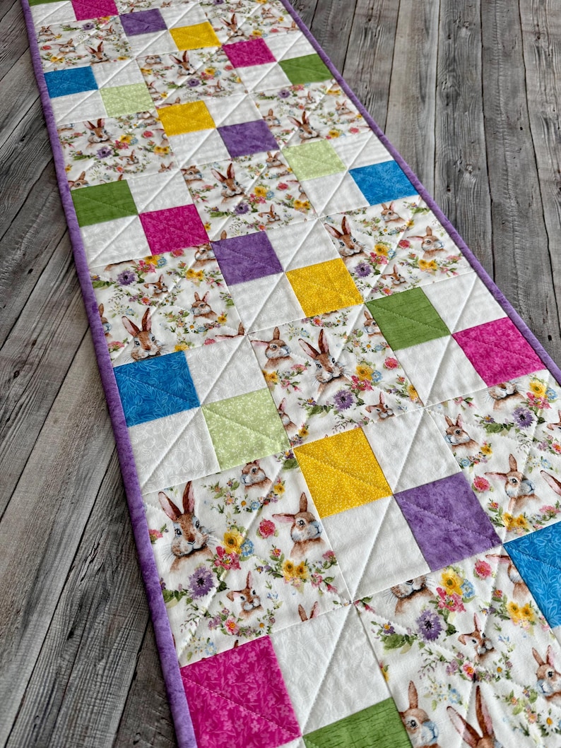 May include: A quilted Easter table runner featuring a repeating pattern of colorful squares and bunny illustrations. The squares are in shades of pink, yellow, green, purple, and blue. The runner has a purple border and is set on a wooden surface.