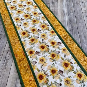May include: A quilted table runner featuring a sunflower and bird pattern on a white background. The runner has a yellow floral border with a green trim. The design is a mix of yellow, brown, and green.