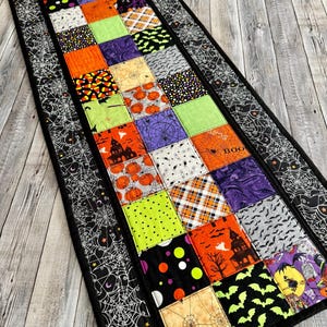 May include: A Halloween-themed table runner with a patchwork design. The runner features various squares with Halloween patterns, including pumpkins, bats, and spiderwebs. The border is black with a white spiderweb design.