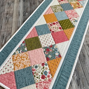 May include: A rectangular quilted table runner featuring a patchwork design of diamond-shaped fabric pieces in various floral, polka dot, and solid patterns in shades of pink, yellow, green, and blue, bordered by a solid blue fabric trim.