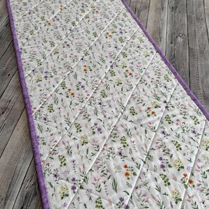 May include: A quilted table runner with a white background, covered in a floral pattern of purple, orange, and green flowers. The edges are trimmed with a solid purple border. The runner is approximately 60 inches long.