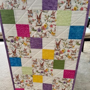 May include: A quilted table runner featuring a patchwork design with squares of white, floral, and solid colors. The floral fabric has bunnies and flowers. The squares are in shades of purple, yellow, green, pink, and blue. The runner is trimmed with a purple border.