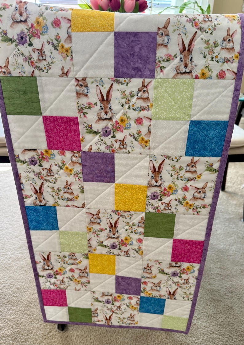 May include: A quilted table runner featuring a patchwork design with squares of white, floral, and solid colors. The floral fabric has bunnies and flowers. The squares are in shades of purple, yellow, green, pink, and blue. The runner is trimmed with a purple border.
