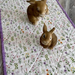 May include: A white floral quilted table runner with a purple border, featuring a pattern of colorful flowers. Two brown rabbit figurines are placed on the runner, creating a spring or Easter theme.