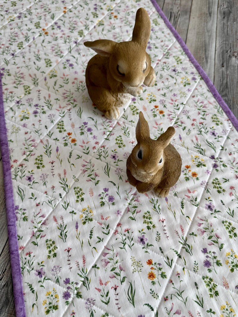 May include: A white floral quilted table runner with a purple border, featuring a pattern of colorful flowers. Two brown rabbit figurines are placed on the runner, creating a spring or Easter theme.