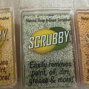 May include: Three natural soap infused scrubbers with the brand name "SCRUBBY" printed on them. Each scrubber features a different fruit: a lemon, a lime, and an orange. The scrubbers are packaged in clear plastic and have a honeycomb-like texture. The text "Easily removes paint, oil, dirt, grease & more!" is printed on each scrubber.