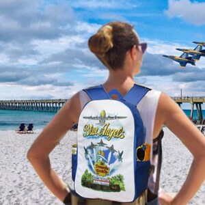 Pu&ograve; includere: Uno zaino blu e bianco con una grafica con la scritta "Blue Angels" e immagini di aerei. Lo zaino &egrave; indossato su una spiaggia con un molo sullo sfondo e le Blue Angels che volano sopra.